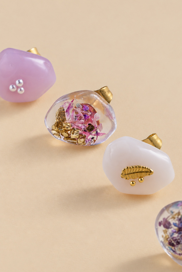 Four resin stud earrings with gold post backs, displayed on a warm beige background. The earrings feature organic shapes with embedded details: one is lavender with silver beads, one is clear with pink dried flowers and gold foil, one is milky white with a gold leaf, and another partially visible one contains purple dried flowers.